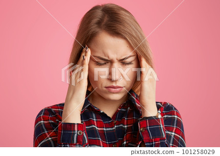 Headshot of stressful woman keeps hands on temples, closes eyes from pain, suffers from migraine, wears stylish shirt, models against pink background, has bad symptoms. Headache and tiredness 101258029