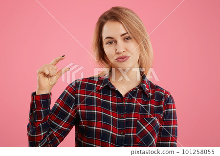Glad positive female with appealing look shows something tiny with fingers, dressed in checkered shirt, isolated over pink background. Adorable woman shows amount of money she has, gestures indoor Glad positive female with appealing look shows something tiny with fingers, dressed in checkered shirt, isolated over pink background. Adorable woman shows amount of money she has, gestures indoor 101258055