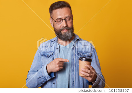 Photo of attractive man pointing on cup of coffee or tea, looking at disponsable mug, posing isolated over yellow background, handsome male with coffee cup dresses jacket. People lifestyle concept. Photo of attractive man pointing on cup of coffee or tea, looking at disponsable mug, posing isolated over yellow background, handsome male with coffee cup dresses jacket. People lifestyle concept. 101258307