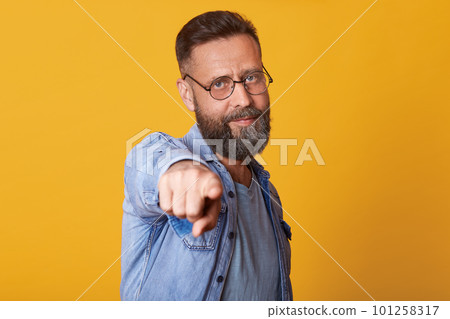 Indoor studio portrait of confident ambitious successful middle aged man posing isolated over bright yellow background, looking directly at camera, showing direction with one forefinger, looks glad. 101258317