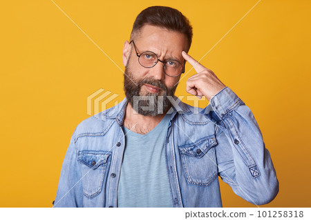 Close up portrait of smart handsome middle aged Caucasian man thinks with finger on head isolated on yellow background, wears denim jacket, gray t shirt, has pansive facial expression. People concept. 101258318