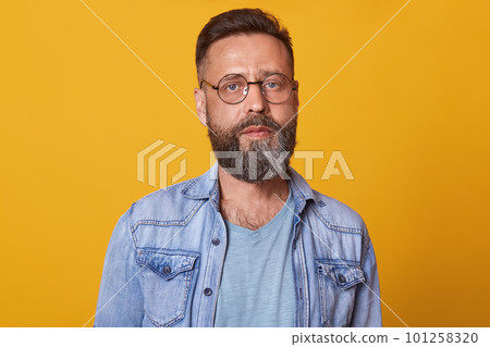 Photo of bearded man with folded hands, standing and looking directly at camera with concentrated facial expression, being in good mood, dressed stylish denim jacket and gray t shirt. People concept. Photo of bearded man with folded hands, standing and looking directly at camera with concentrated facial expression, being in good mood, dressed stylish denim jacket and gray t shirt. People concept. 101258320