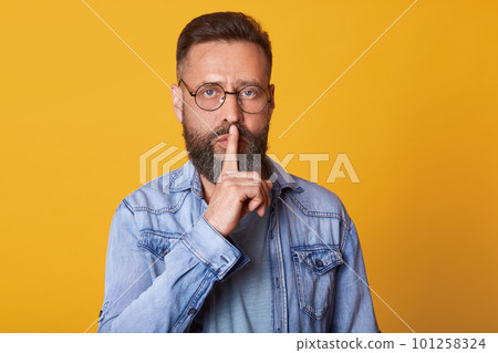 Serious middle aged emotionless bearded man in blue shirt, showing silence gesture, asks to keep secret, putting forefinger on lips, male dressed in denim jacket stands over pink background. Serious middle aged emotionless bearded man in blue shirt, showing silence gesture, asks to keep secret, putting forefinger on lips, male dressed in denim jacket stands over pink background. 101258324