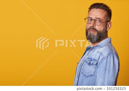 Image of handsome middle aged bearded man with beard, standing and looking directly at camera, posing over yellow background, wearing senim jacket. Copy space for advertismant or promotion text. Image of handsome middle aged bearded man with beard, standing and looking directly at camera, posing over yellow background, wearing senim jacket. Copy space for advertismant or promotion text. 101258325