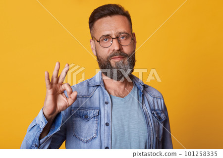 Studio shot of handsome man with beard wearing denim jacket, gray t shirt and spectacles, has positive facial expression, doing ok sign with hand and fingers, isolated over yellow studio background. 101258335