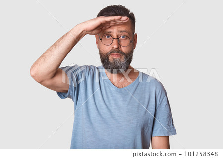 Half length studio shot of handsome male posing isolated over white studio background, peeks out someone, keeps hand near forehead, man dressed casually, has round spectacles, with serious expression. 101258348