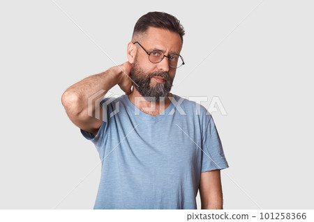 Tired unemotional handsome executive looks firmly at camera, holding his arm behind, making neck massage after hard working day. Middle aged black haired model wearing stylish specs and t shirt. Tired unemotional handsome executive looks firmly at camera, holding his arm behind, making neck massage after hard working day. Middle aged black haired model wearing stylish specs and t shirt. 101258366
