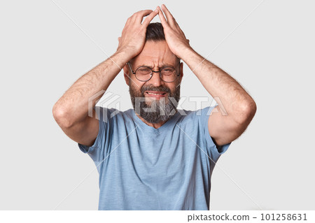 Bearded man in gray t shirt, has round eyewear, keeps hand on head, grimaced, has problems, being in bad mood, has terrible headache, being in conflict wit his wife. People emocions concept. Bearded man in gray t shirt, has round eyewear, keeps hand on head, grimaced, has problems, being in bad mood, has terrible headache, being in conflict wit his wife. People emocions concept. 101258631