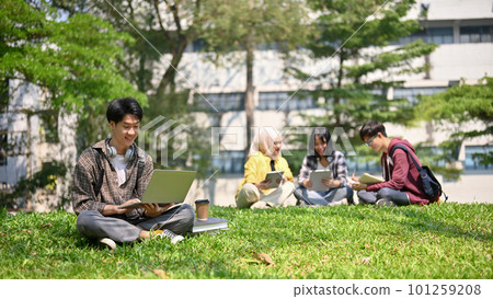 Handsome Asian male college student sits on the grass in the campus park and uses his laptop Handsome Asian male college student sits on the grass in the campus park and uses his laptop 101259208