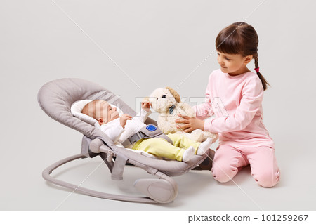 Happy sibling posing together isolated over white background, elder sister showing soft dog toy to little cute baby lying in rocking chair, happy childhood. 101259267