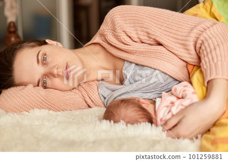 Soft photo of young mother feeding breast her baby at home while lying in bed, mommy looking at camera with tired and sleepless facial expression, wearing home clothing. Soft photo of young mother feeding breast her baby at home while lying in bed, mommy looking at camera with tired and sleepless facial expression, wearing home clothing. 101259881