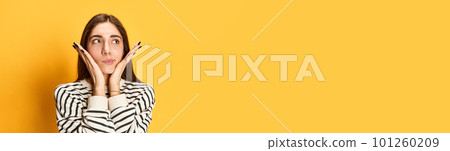 Portrait of young girl in stripped sweater holding palms near face and thinking, posing agaisnt yellow studio background 101260209