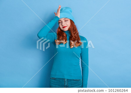 Young attractive female with red hair posing against blue background in cap and warm sweater, touching her forehead with palm, forget about important thing, touches her forehead. 101260264