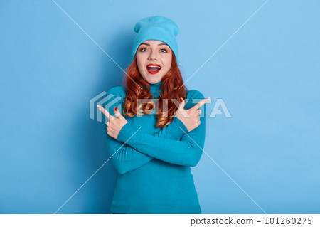 Good looking female isolated over blue background, pointing with both hands to both sides, keeping mouth open wide, being surprised and astonished about excited news, wearing jumper and cap. 101260275