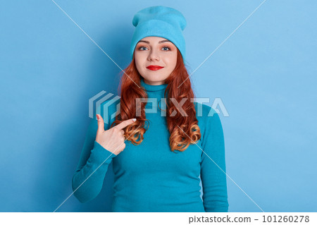 Satisfied Caucasian woman with red hair points at herself, stands self confident, dressed casual sweater and cap, feels successful of her own achievement, stands indoor against blue background. 101260278
