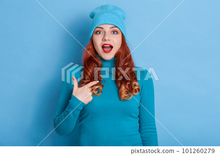 Indoor shot of cheerful smiling young Caucasian woman with curls pointing index finger on her self, keeps mouth opened, posing with big eyes, wearing casually, isolated over blue background. 101260279
