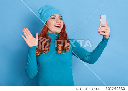 Excited female wearing blue jumper and cap, posing with raising palm, having video call or taking selfie, looks happy, wears red lipstick, has curls, holds smart phone in hand. 101260280