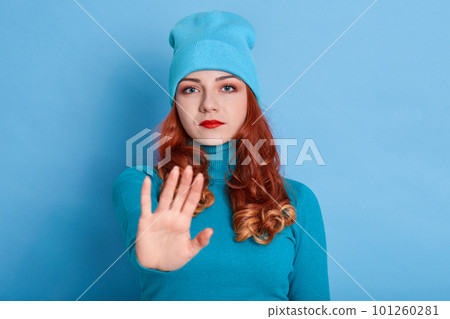 Confident redhead woman keeps palms towards camera, has displeased expression, looks at camera, makes stop gesture, ginger lady gives warning prohibits action, wearing blue sweater and cap. 101260281