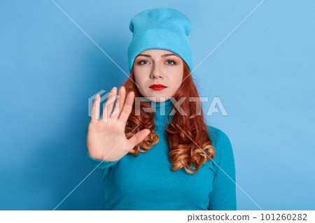 Lady making stop gesture with her palm, posing over blue background, looks strict and serious, wearing sweater and cap, has red wavy hair, something forbids. Lady making stop gesture with her palm, posing over blue background, looks strict and serious, wearing sweater and cap, has red wavy hair, something forbids. 101260282