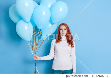 Unhappy sad young woman with balloons looking at camera, lady with red hair, posing against blue background, female being offended, expresses negative emotions. Unhappy sad young woman with balloons looking at camera, lady with red hair, posing against blue background, female being offended, expresses negative emotions. 101260297