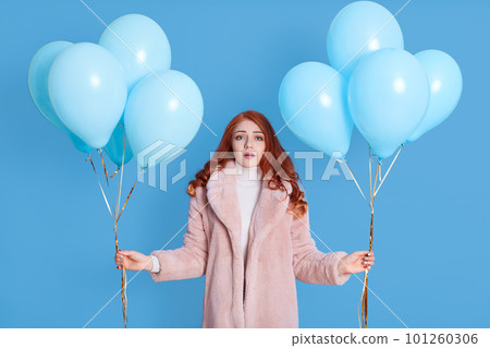 Young caucasian woman looks scared and shocked, looks at camera with opened mouth, being astonished, holding bunches with helium balloons in hands, dresses pale pink fur coat, posing against blue wall 101260306