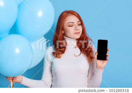 Pensive red haired European woman thinks celebrate birthday holiday party, holds modern mobile phone with blank screen and bunch of helium balloons in hands, wears white turtleneck, female looks away. 101260318