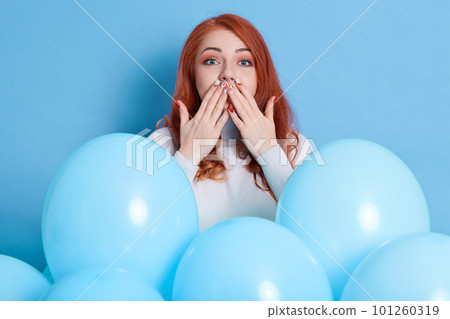 Pretty red haired girl wearing casually, covering mouth with fingers while looking directly at camera, being surrounded with balloons posing isolated over blue background. 101260319
