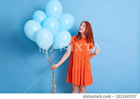 Woman holding bunch of helium balloons, looking at her present with surprised and excited facial expression, standing isolated over blue background. Woman holding bunch of helium balloons, looking at her present with surprised and excited facial expression, standing isolated over blue background. 101260337