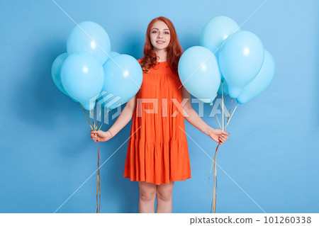 Young pretty woman with two bunches of balloons standing against blue wall and looking smiling directly at camera, celebrating holiday, girl with red hair wearing dress . Young pretty woman with two bunches of balloons standing against blue wall and looking smiling directly at camera, celebrating holiday, girl with red hair wearing dress . 101260338
