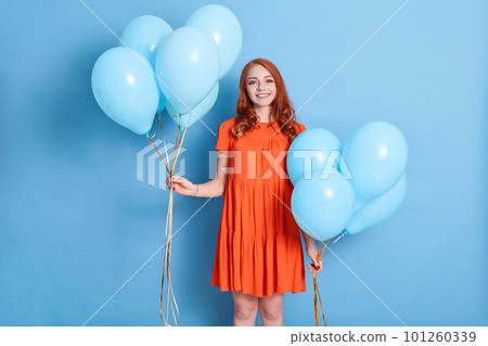 Pretty woman with balloons, wearing elegant dress, has red wavy hair, looks at camera, happy woman celebrating her birthday, standing isolated over blue background. Pretty woman with balloons, wearing elegant dress, has red wavy hair, looks at camera, happy woman celebrating her birthday, standing isolated over blue background. 101260339