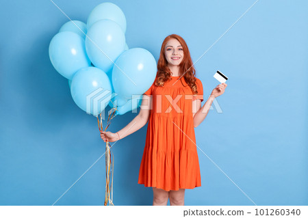 Cheerful pretty young woman holding credit bank card, celebrating with helium balloons isolated over blue background. Birthday holiday party, wearing dress. Cheerful pretty young woman holding credit bank card, celebrating with helium balloons isolated over blue background. Birthday holiday party, wearing dress. 101260340