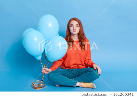 Winsome girl dressed in casual shirt and jeans holds balloons in her hands while sitting on floor isolated over blue background, looking at camera. Winsome girl dressed in casual shirt and jeans holds balloons in her hands while sitting on floor isolated over blue background, looking at camera. 101260341
