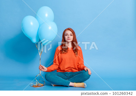 Ginger European girl dressed in orange jumper and jeans holds balloons in her hands while sitting on floor with crossed legs, red haired lady has birthday, poses on blue background. Ginger European girl dressed in orange jumper and jeans holds balloons in her hands while sitting on floor with crossed legs, red haired lady has birthday, poses on blue background. 101260342