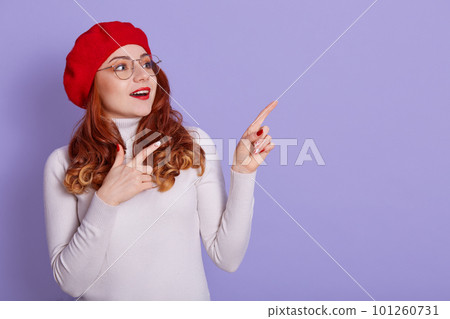 Cute happy woman gives direction, points with both hands aside, has overjoyed facial expression, wears beret, white casual shirt and spectacles, models over lilac background. 101260731