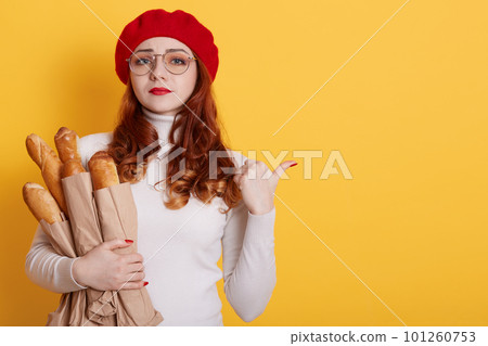 Young beautiful upset woman holding bag of fresh healthy bread over yellow background,red haired sad girl indicating aside, looks worried and nervous, lady wearing beret, white shirt and glasses. Young beautiful upset woman holding bag of fresh healthy bread over yellow background,red haired sad girl indicating aside, looks worried and nervous, lady wearing beret, white shirt and glasses. 101260753
