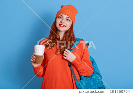 Image of joyful caucasian girl in hat drinking coffee while holding back pack, looks at camera, ginger lady has break between lectures, smiling girl enjoying hot take away coffee. Image of joyful caucasian girl in hat drinking coffee while holding back pack, looks at camera, ginger lady has break between lectures, smiling girl enjoying hot take away coffee. 101260794