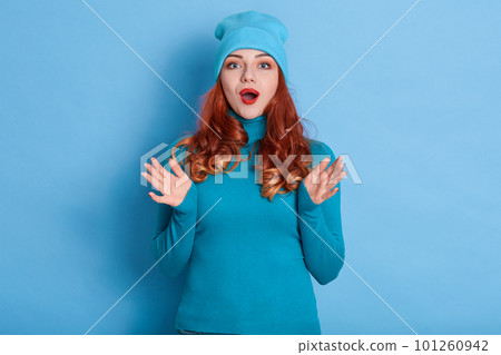 Closeup portrait of shocked girl with big beautiful eyes and red lipstick, looks at camera with his palms spreading, posing isolated over blue background, wearing casually. 101260942