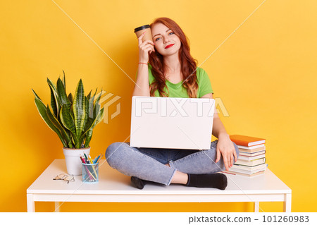 Girl student sitting with cup of coffee in hand, holds beverage near temple, being tired but happy, looks at camera with smile, girl wearing casually, sits on table near flower pot, stack of books. Girl student sitting with cup of coffee in hand, holds beverage near temple, being tired but happy, looks at camera with smile, girl wearing casually, sits on table near flower pot, stack of books. 101260983