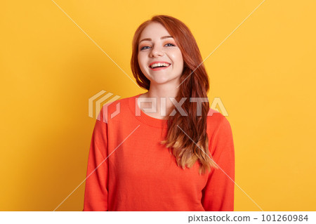 Laughing woman with wavy hair isolated on yellow wall looks at camera and expressing happiness, wearing orange sweater, smiling happily, ginger satisfied girl in good mood. 101260984