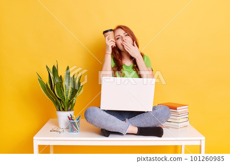 Exhausted European woman with red hair being tired of university and diploma, sitting with laptop on knees, yawning and covering mouth with palm, keeping disposable cup of coffee near head. 101260985