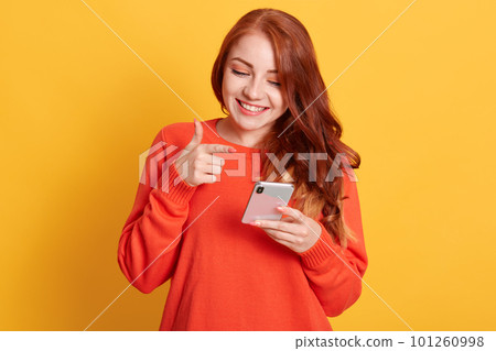 Cheerful woman pointing to her smart phone screen with index finger, looking at mobile phone with charming smile, standing isolated over yellow background. 101260998