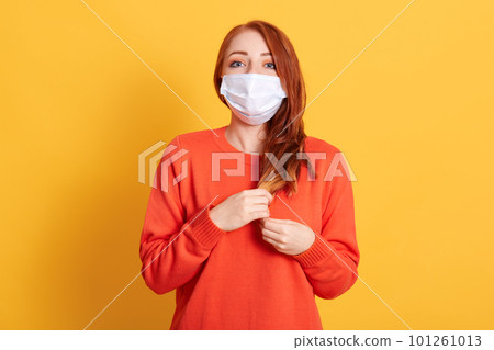 Gorgeous red haired young female wearing protective mask against COVID virus, tired of stress, looks confidently at camera with fear in eyes, poses against yellow background. Gorgeous red haired young female wearing protective mask against COVID virus, tired of stress, looks confidently at camera with fear in eyes, poses against yellow background. 101261013