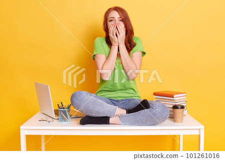 Young shocked woman in casual clothes covering mouth with hands while sitting on desk with crossed legs, surrounded with pc laptop, books, coffee isolated on yellow background. 101261016