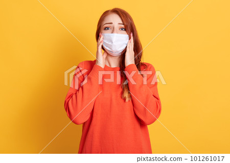 Portrait of young red haired girl with beautiful eyes, having headache, holding hands on cheeks, wearing medical flu mask and orange sweater isolated over yellow background. 101261017