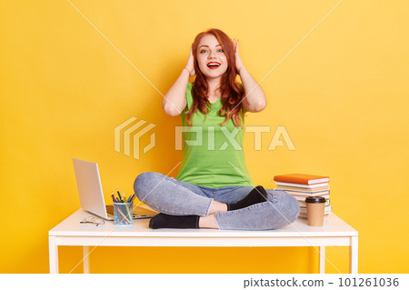 Red haired girl pressing her hands to her head, looks at camera with opened mouth and excited facial expression, wearing green t shirt and jeans, posing on desk with crossed legs. Red haired girl pressing her hands to her head, looks at camera with opened mouth and excited facial expression, wearing green t shirt and jeans, posing on desk with crossed legs. 101261036