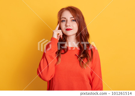 Young adult girl posing with pensive look, holding finger on temple. Attractive woman in orange sweater having new great idea, looks at camera, posing isolated over yellow wall. 101261514