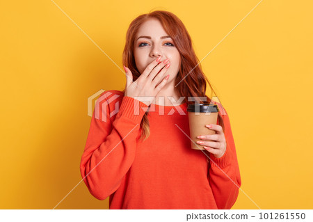 Exhausted sleepy female has sleepless night of exam preparation, yawns and covers mouth with palm drinks takeaway coffee to feel refreshed, red haired lady wearing orange sweater. Exhausted sleepy female has sleepless night of exam preparation, yawns and covers mouth with palm drinks takeaway coffee to feel refreshed, red haired lady wearing orange sweater. 101261550