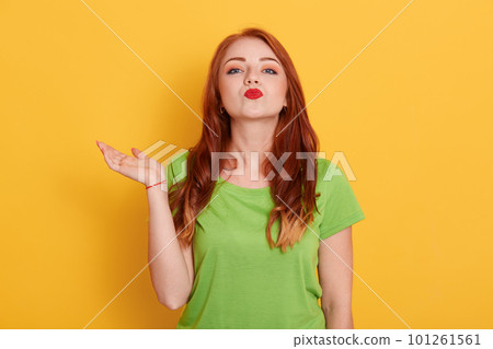 Smiling European female with charming facial expression wearing green casual t shirt, spreading palm aside, looks at camera and making kiss gesture, standing isolated over yellow background. 101261561