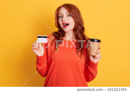 Astonished buyer finding offer online, holding take away coffee and credit card, has surprised facial expression, lady with red lips and wavy hair, wearing orange sweater. Astonished buyer finding offer online, holding take away coffee and credit card, has surprised facial expression, lady with red lips and wavy hair, wearing orange sweater. 101261565
