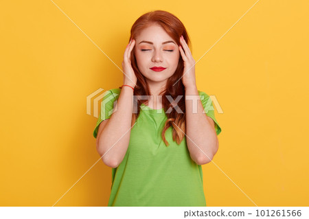 Portrait of charming, stylish woman in green shirt having head ache, touching temples with fingers and close eyes, standing over yellow background. 101261566